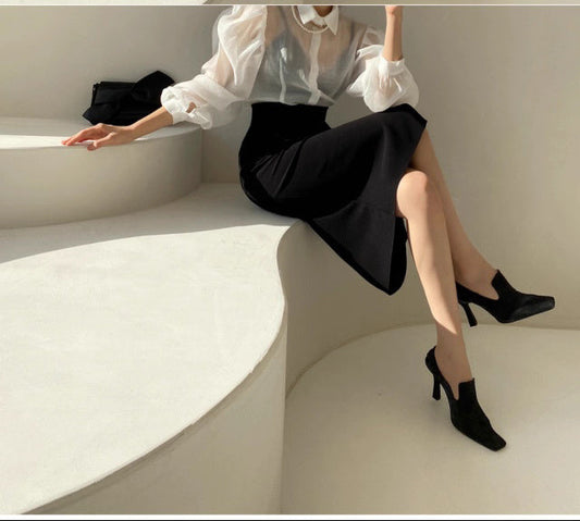 Woman wearing a chic sheer office blouse paired with a black skirt and black heels sitting on a curved white bench