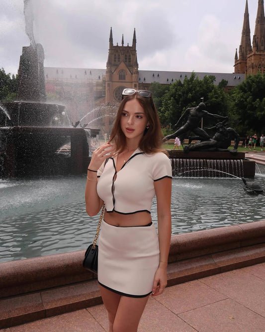 Woman wearing Orchid Moda beige desert co-ord set with black trim standing by a city fountain