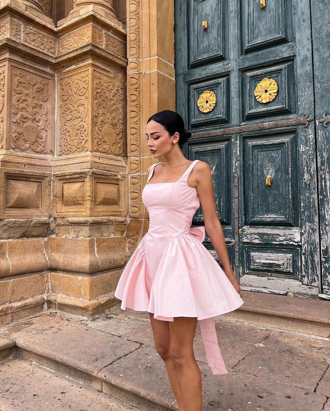 Woman wearing a pink knotted bow mini dress for women standing by ornate stone building and wooden door