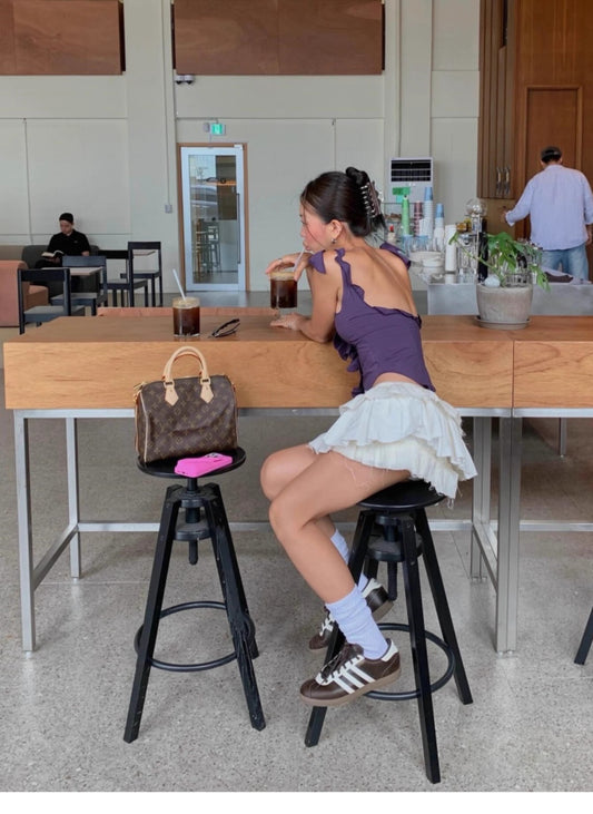Woman wearing ruffled cami top for women in purple paired with white ruffled skirt sitting on stool in cafe