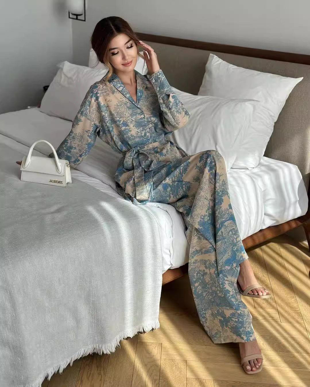Woman wearing floral patterned women's co ord set sitting on bed with white pillows and purse